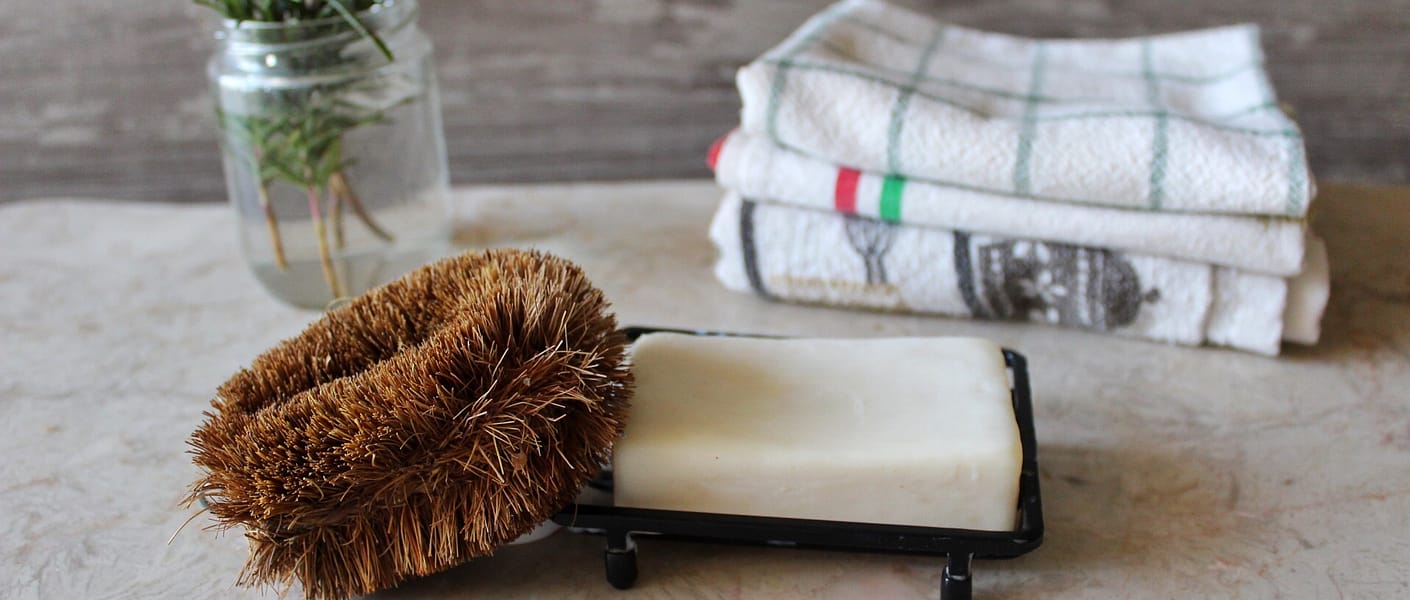 bar of soap with brown dish scrubber leaning against it, jar of herbs and stacked towels in background