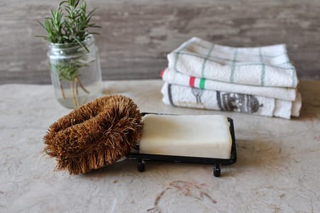 bar of soap with brown dish scrubber leaning against it, jar of herbs and stacked towels in background